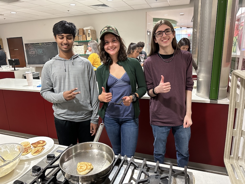 Three students give thumbs up and smile in front of a stove and frying pan for pancakes