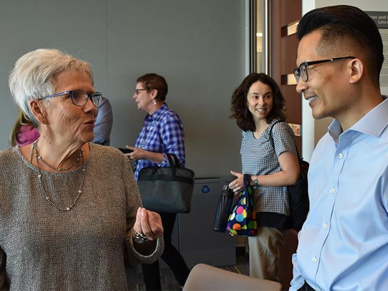 Two faculty and staff and MLFC smile and talk with each other, as others in the background file into a conference room for a meeting