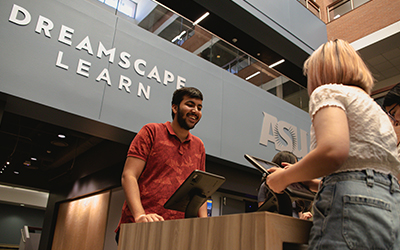 A student standing in front of a check-in station smiles at another, in front of a sign that reads Dreamscape Learn