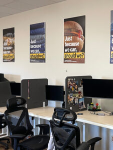 Posters containing the text "Just because we can, should we?" over images of food in plastic containers, a pile of blue jeans, and a hamburger, hang on the wall over a row of computer stations.