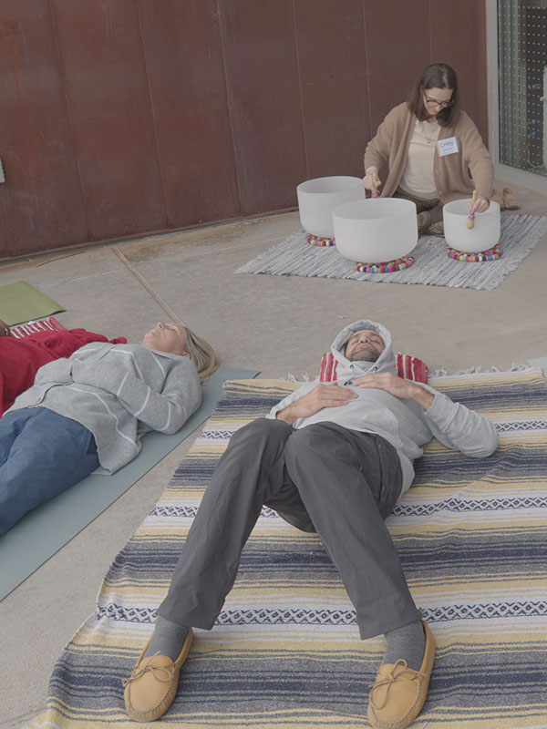 Two people lay on carpets on their backs as a seated woman plays crystal singing bowls